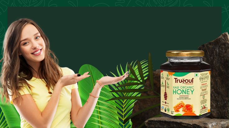 Smiling woman in a yellow shirt presenting a jar of TruSoul Raw Organic Honey, placed on a rock with lush green leaves in the background.