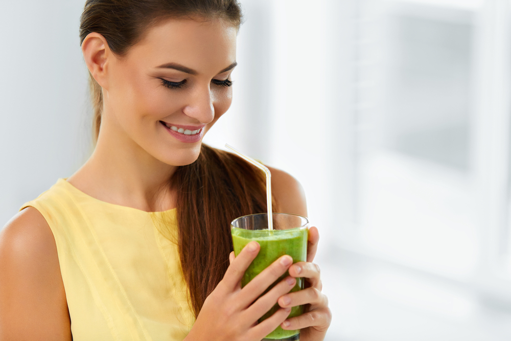 Young woman smiling while drinking green juice through a straw, promoting healthy lifestyle and natural wellness.